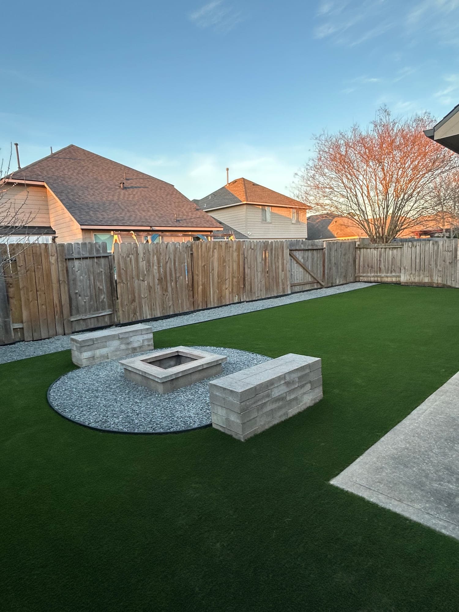 The same backyard finished with artificial turf, a gravel ring, a squared fire pit and matching block benches at golden hour.