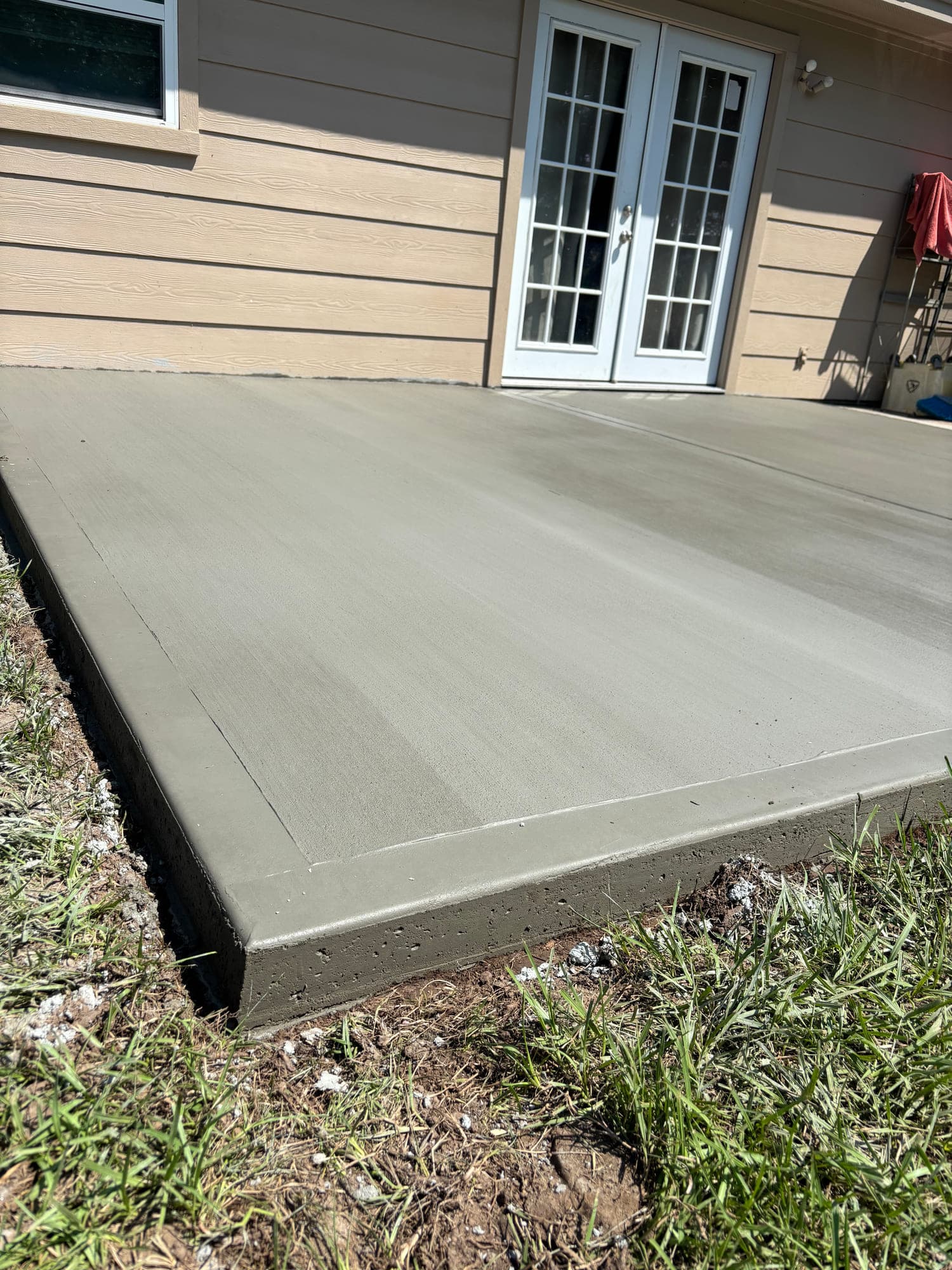Freshly poured concrete patio slab with a clean broom finish and crisp chamfered edge next to french doors.