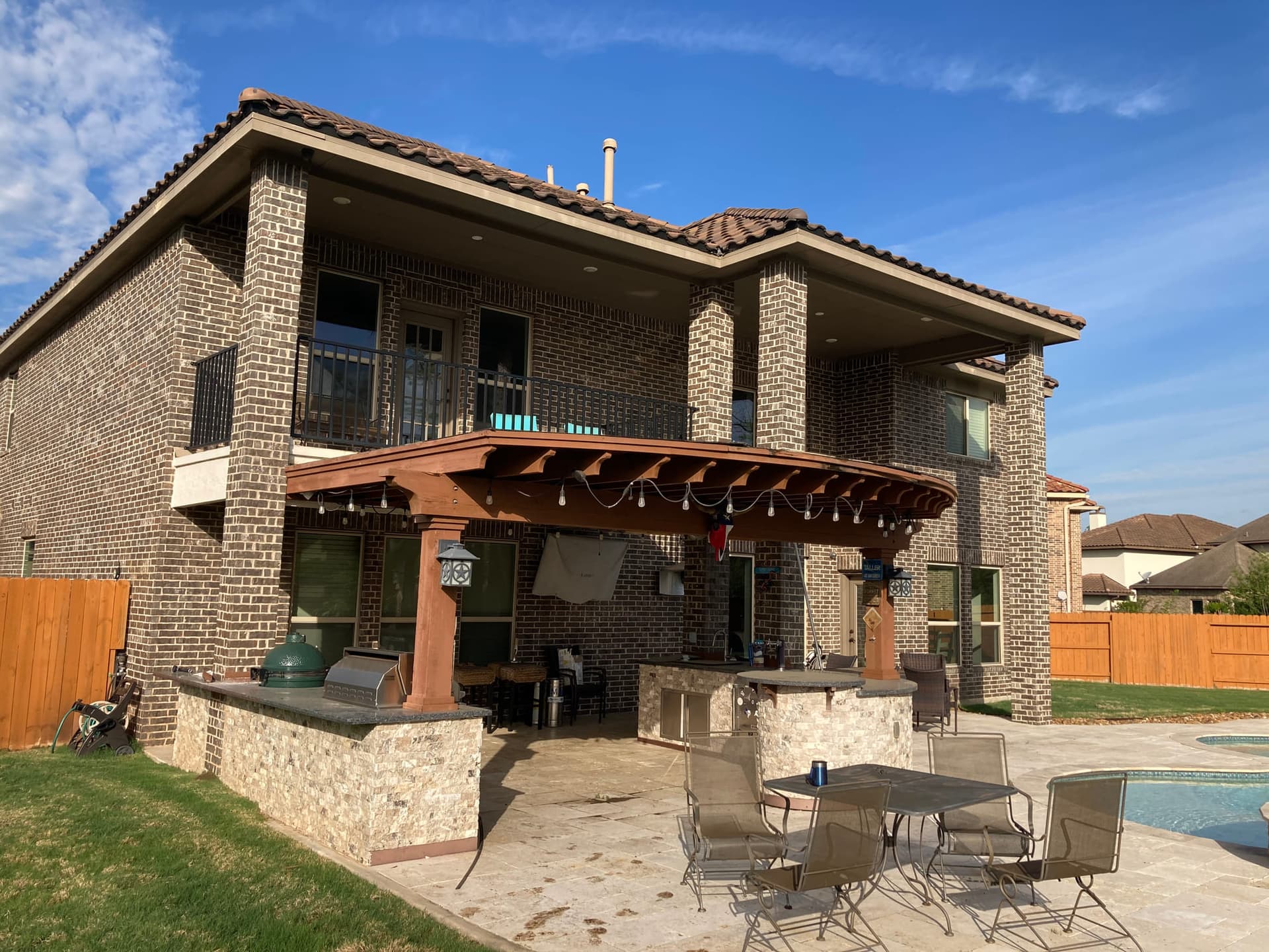 Curved stained-cedar pergola with string lights over a stone outdoor kitchen and travertine pool deck behind a two-story brick home.
