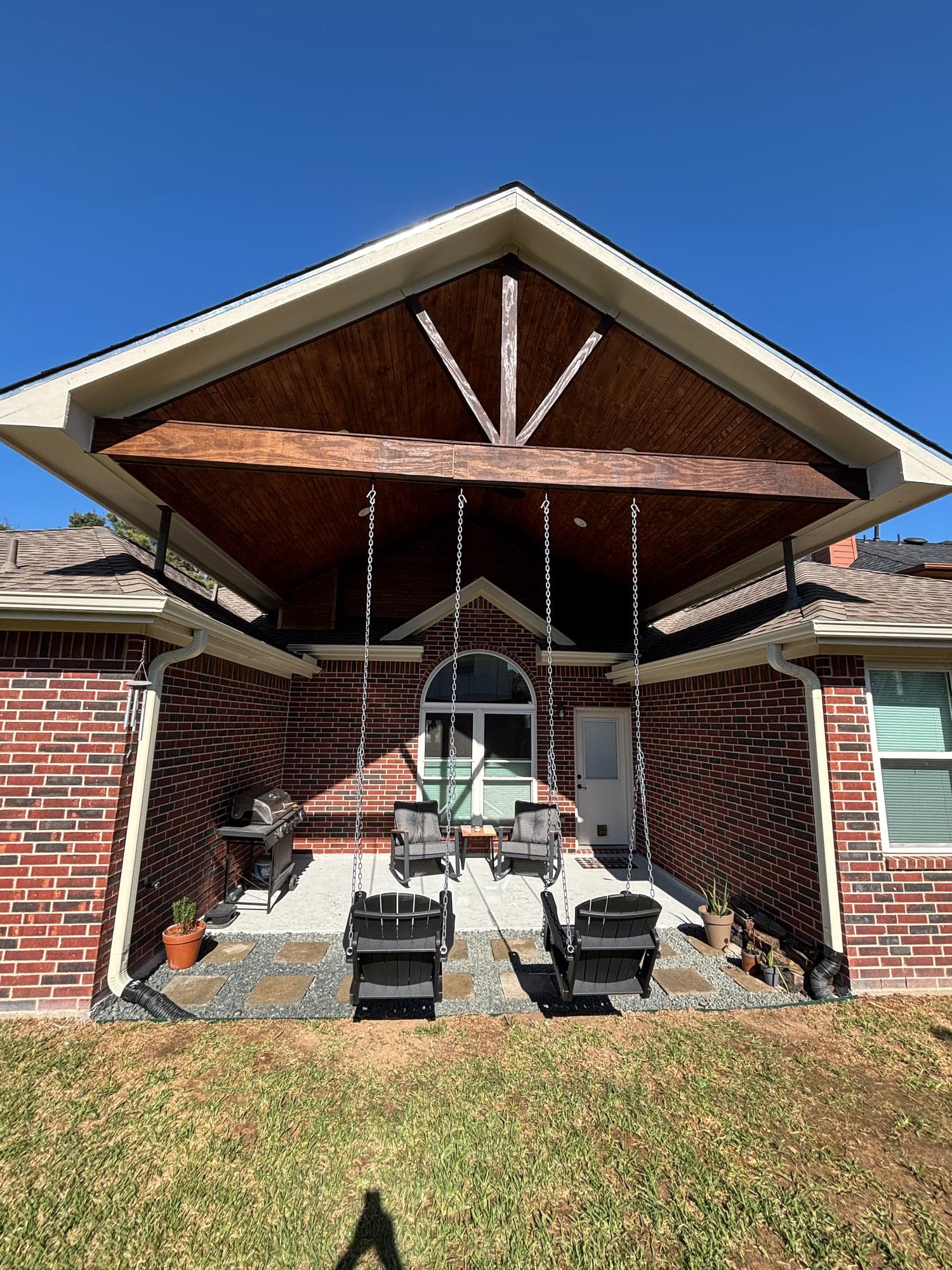 Open-gable patio cover with a stained wood ceiling and two hanging porch swings over a concrete and flagstone patio.